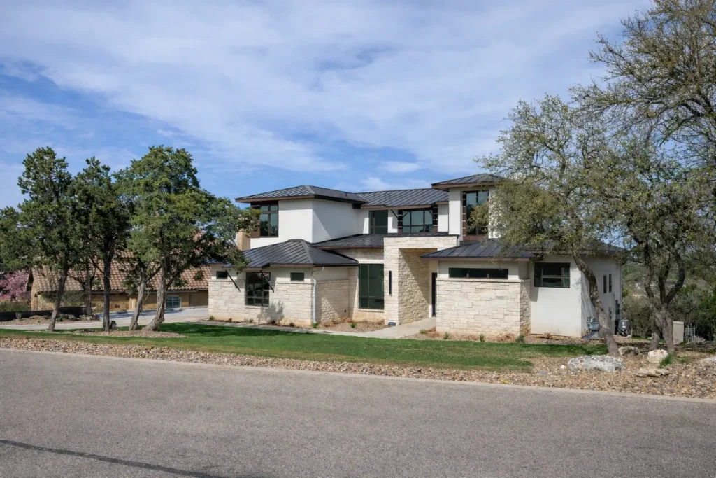 Street view of a two-story limestone and stucco contemporary custom home with a metal roof and mature oaks