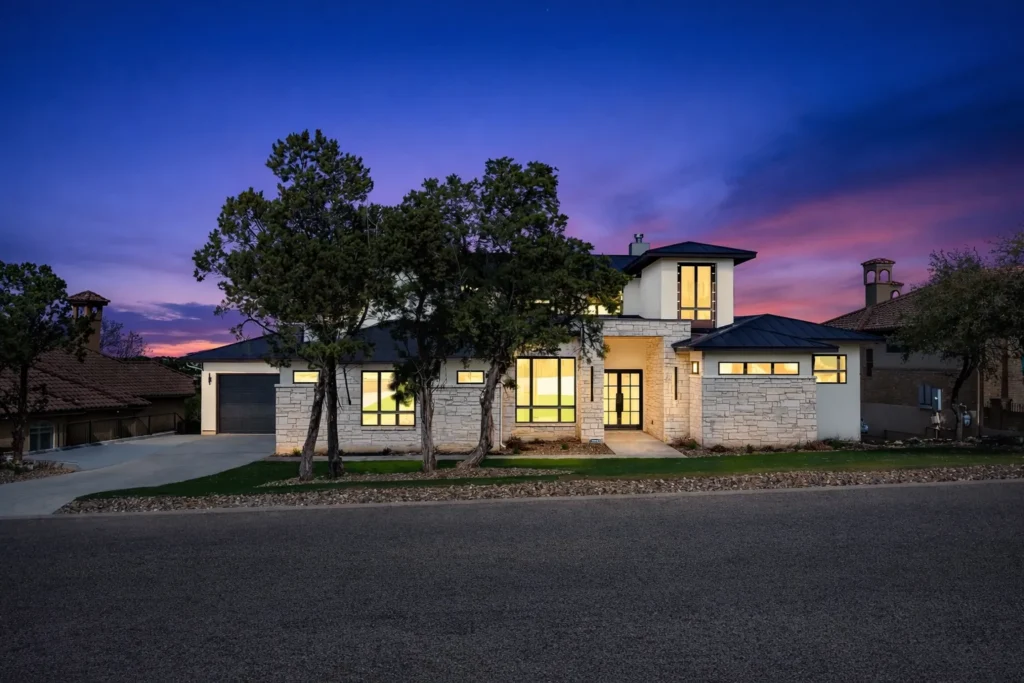 Twilight exterior of a two-story Hill Country contemporary custom home with limestone walls, a metal roof and glowing windows