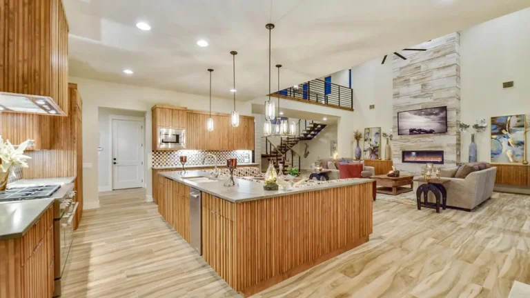 Open kitchen with a slatted wood island, pendant lights and wood cabinetry facing a great room with a limestone fireplace and catwalk