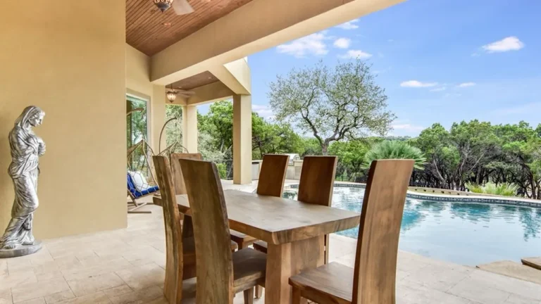 Covered patio with a solid-wood dining table, statue and hanging chair beside a pool overlooking Hill Country oaks