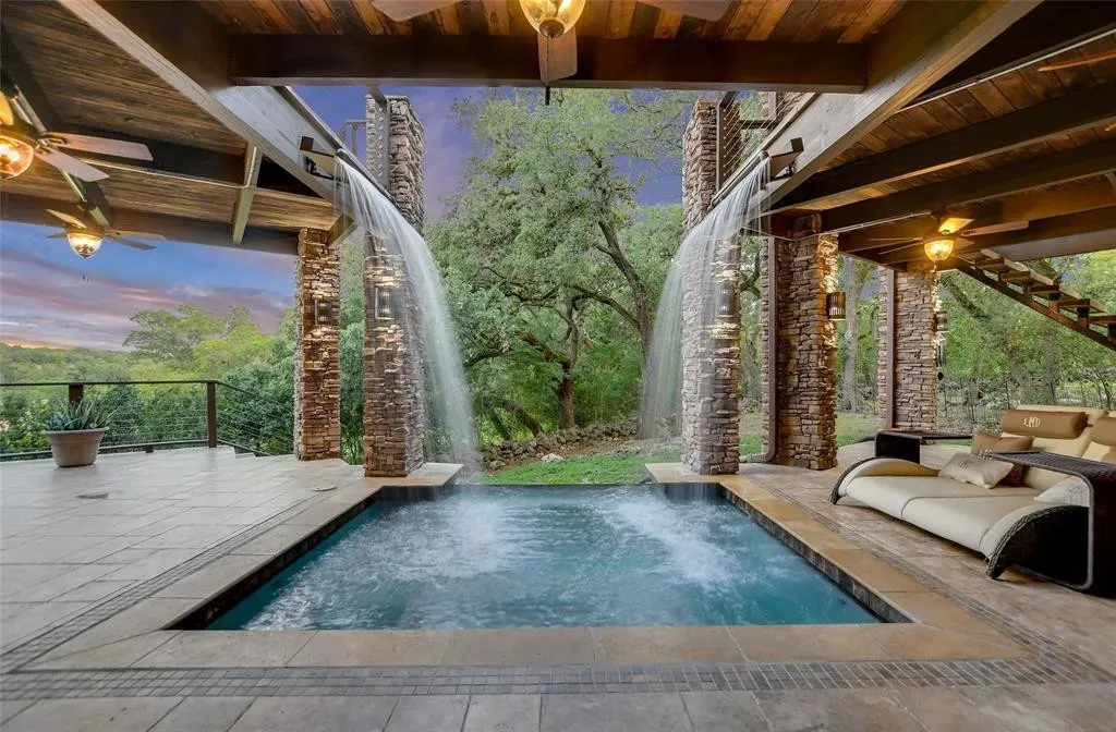 Covered patio with twin stone waterfall columns spilling into a plunge pool beneath a cedar ceiling, overlooking Hill Country oaks