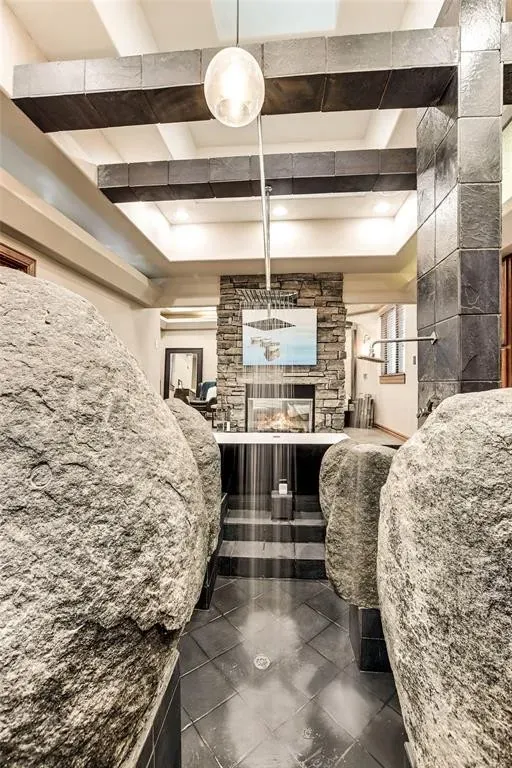Primary bath entry framed by natural boulders leading to a stone shower and soaking tub under dark timber beams