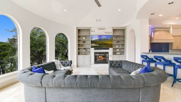 Family room with a gray tufted sectional, fireplace and built-ins framed by arched windows, open to a kitchen with blue bar stools