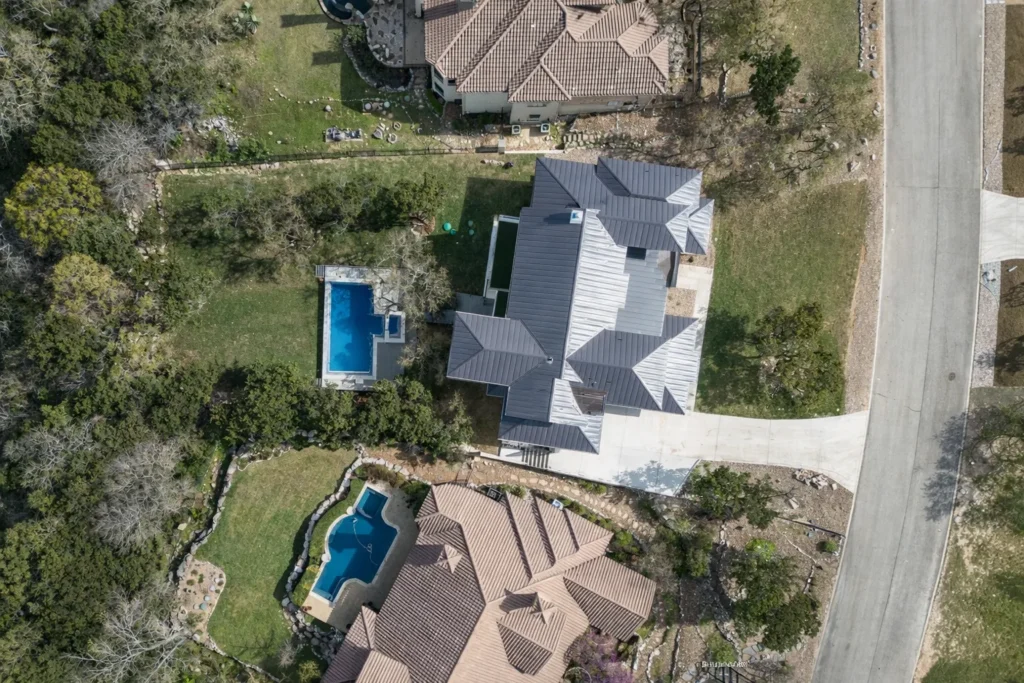 Overhead aerial view of a custom home with a metal roof, rectangular pool and landscaped Hill Country lot