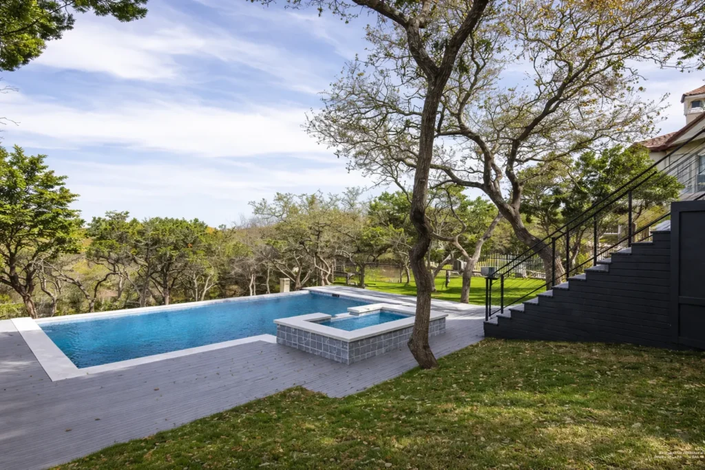 Rectangular pool and raised spa on a gray deck shaded by live oaks behind a modern custom home