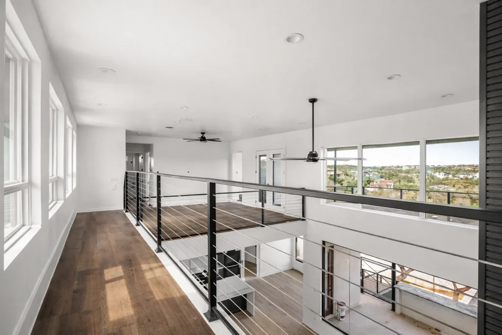 Upstairs loft walkway with black cable railings and hardwood floors overlooking the living area and Hill Country views