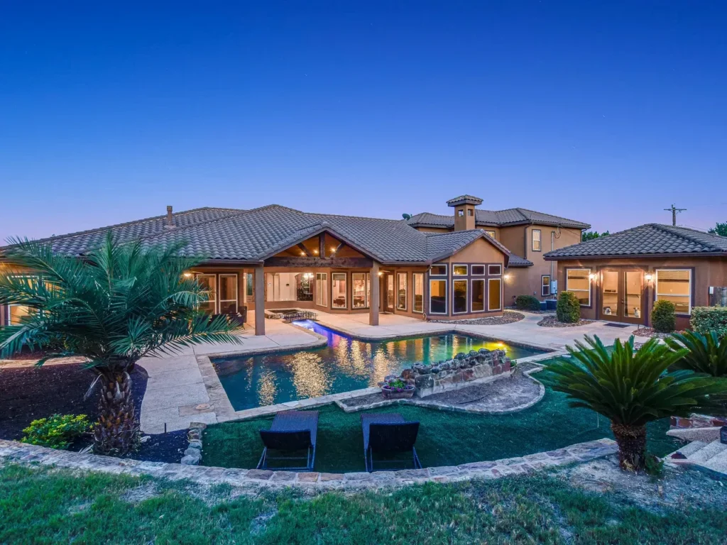 Elevated twilight view of a backyard pool with a rock waterfall, palms and covered patio at a Hill Country estate