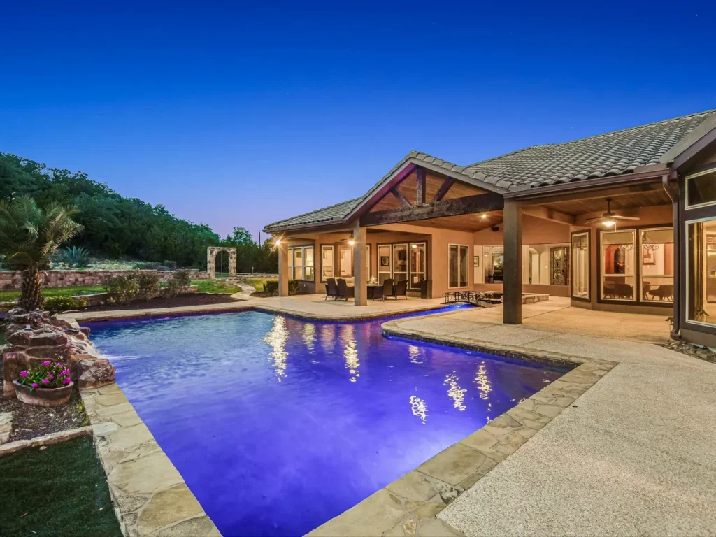 Freeform pool glowing with color lighting beside a stone and stucco home with a timber gable patio at dusk