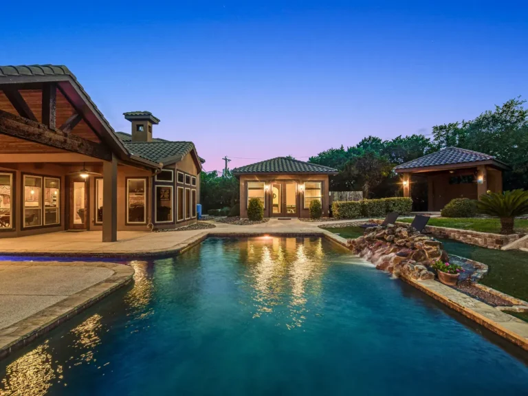 Pool with a natural rock waterfall, pool house casita and covered patio on a wooded Hill Country lot at dusk