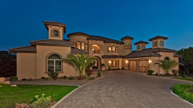 Mediterranean custom home lit at dusk with tower rooms, an arched balcony, carriage-style garage doors and a paver motor court