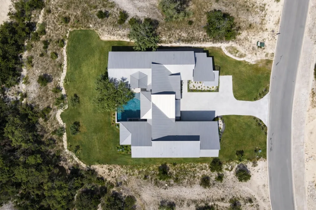 Overhead aerial view of a single-story modern custom home with a metal roof and pool on a wooded Hill Country lot