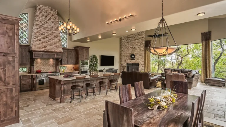 Knotty alder kitchen with a brick range hood, iron chandeliers and island seating open to a stone fireplace family room and dining table