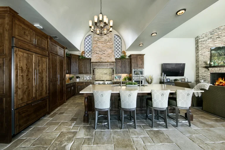 Kitchen island with upholstered stools beneath an iron chandelier, flanked by paneled alder refrigeration and arched windows