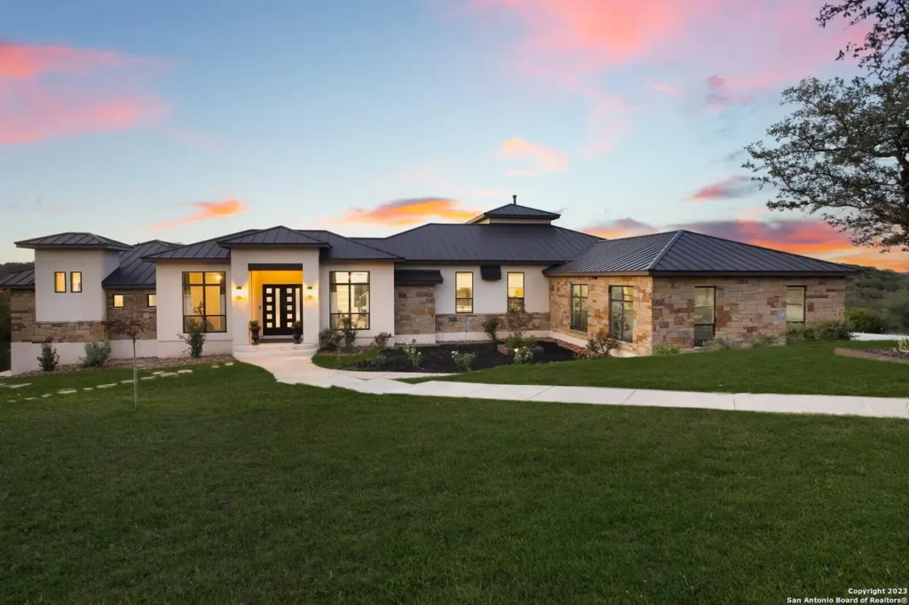 Single-story stone and stucco custom home with a metal roof and glowing entry at sunset in the Texas Hill Country