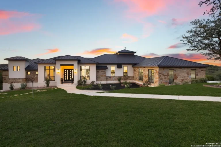 Single-story stone and stucco custom home with a metal roof and glowing entry at sunset in the Texas Hill Country