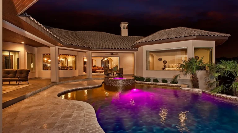 Freeform pool with color lighting, a raised spa and travertine deck wrapped by a covered patio at night