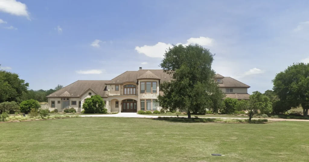 Two-story stone and stucco estate home with arched windows set behind a broad lawn and live oaks in the Texas Hill Country