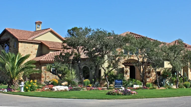 Two-story stone and stucco custom home with a tile roof, chimney and live oaks behind colorful flower beds