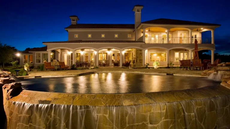 Two-story arched loggias glowing at night above a vanishing-edge pool with a stone waterfall at a custom home in Mystic Shores, Canyon Lake