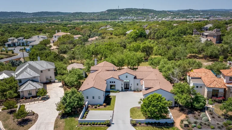 Aerial view of a white stucco custom home with a clay tile roof among wooded homesites and rolling hills in The Dominion, San Antonio