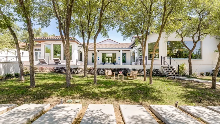 Shaded backyard lawn with live oaks and concrete step pads leading to the rear patios of a white stucco home