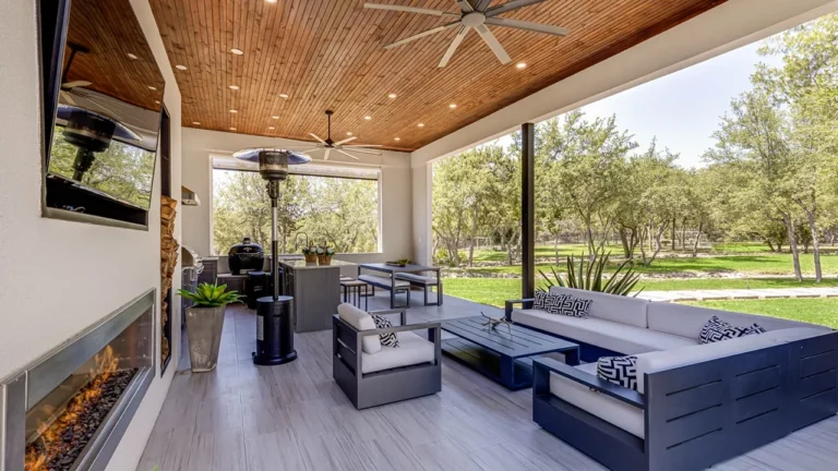 Covered outdoor living room with a linear fireplace, mounted TV, cedar plank ceiling and modern seating overlooking the wooded yard