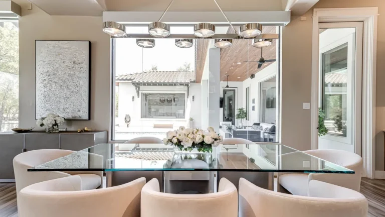 Dining area with a glass table, white swivel chairs and a linear chandelier facing windows to the covered patio