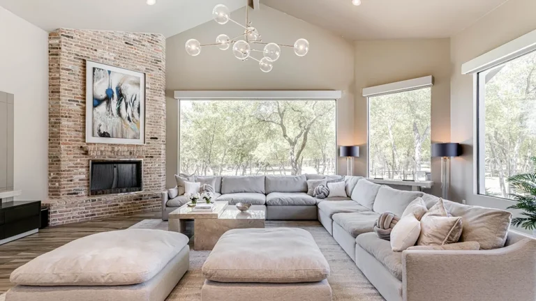 Great room with a brick fireplace wall, oversized white sectional, glass-globe chandelier and picture windows framing live oaks