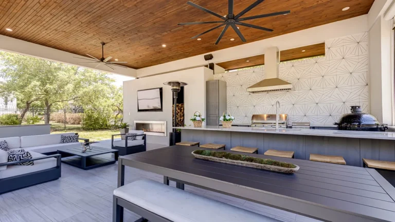 Outdoor kitchen with a geometric white tile backsplash, stainless hood, built-in grill, kamado grill and long dining table under a cedar ceiling
