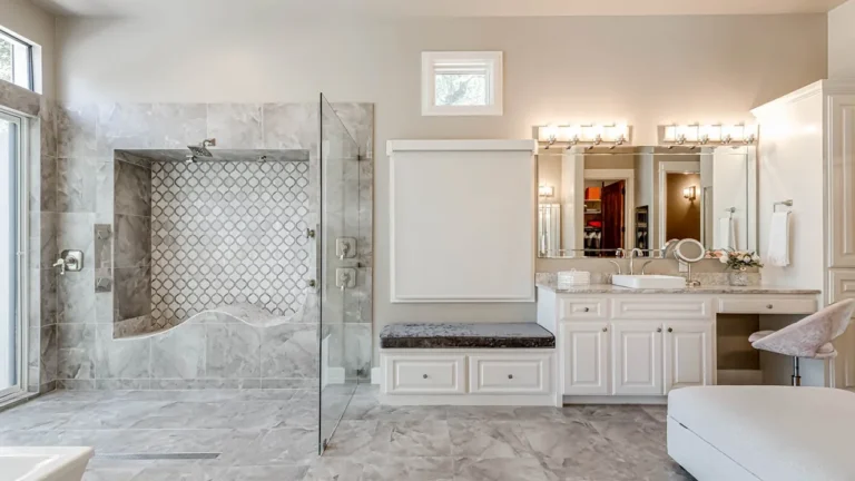 Walk-in glass shower with an arabesque mosaic wall beside white vanities, a tufted bench and a makeup station