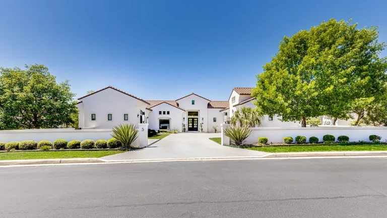 Street view of a white stucco, clay tile roof custom home behind a low garden wall and mature shade trees in The Dominion, San Antonio